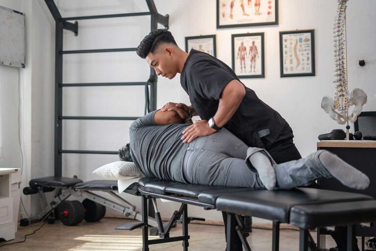 A chiropractor gently adjusting a patient's upper back as the patient lies face down on an exam table.